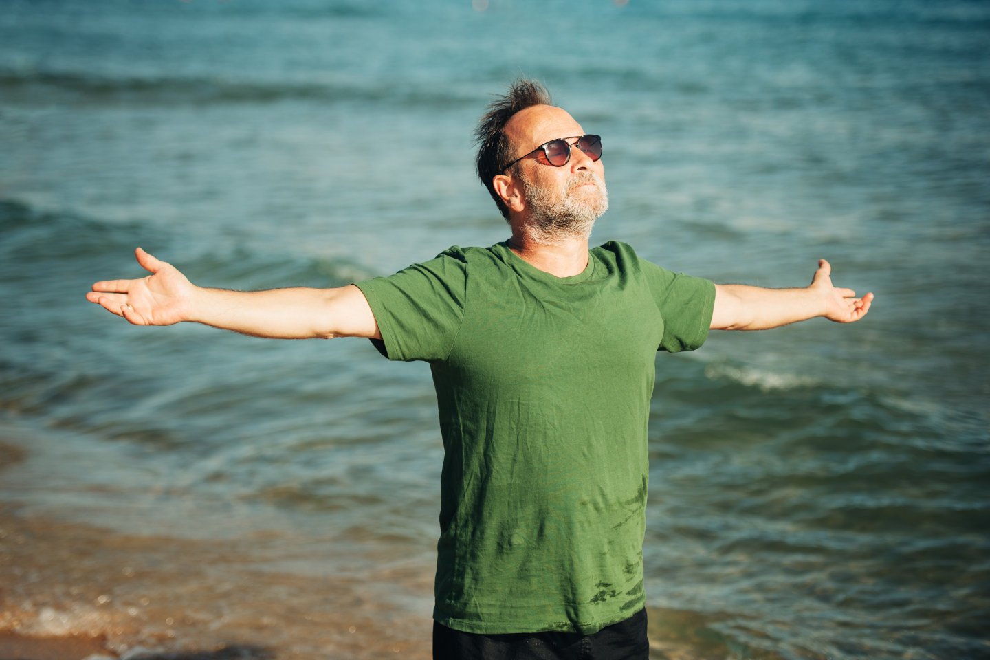 Portrait of happy middle age man enjoying nice sunny day on the beach, wearing green t-shirt and sunglasses, arms wide open